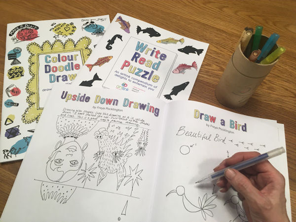 Hand colouring 'Draw a Bird' with 'Colour Doodle Draw' and 'Write Read Puzzle' pamphlets on table.