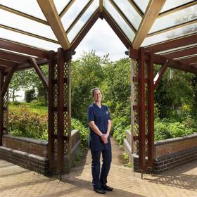 A young woman in a T-shirt stands on a sunny patio, framed by the pillars of an attractive wooden conservatory, greenery and flowerbeds behind her.