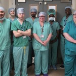 Eight healthcare staff in green scrubs stand in a hospital corridor outside theatre doors.