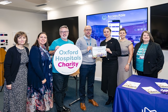 Seven people in front of a screen that says 'AccessAble'. Two people hold a plaque that celebrates the launch of the new guides. Two people wear 'Oxford Hospitals Charity' branded t-shirts, while one holds a banner that has the same name.