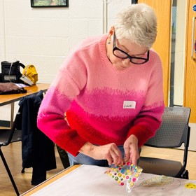 An older woman with a 'Julie' name sticker puts small decorative stickers around a circular drawing on the corner of a sheet.