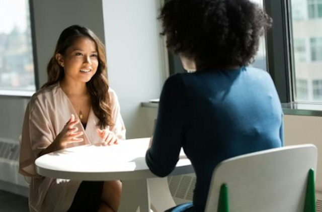 Two women speak to each other across a table in an office setting
