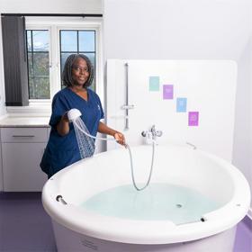 A clean modern room with a large round birthing pool, which a young woman in scrubs is filling with a showerhead. A frosted window behind lets in natural light.