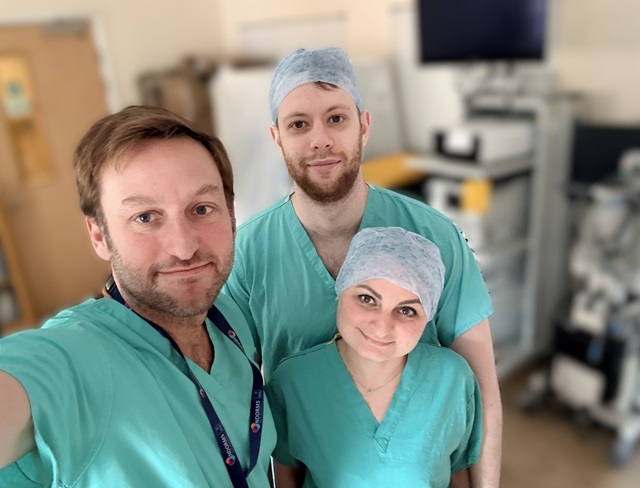 Three healthcare professionals wearing green surgical scrubs in a clinical setting, with medical equipment in the background.