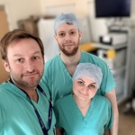 Three healthcare professionals wearing green surgical scrubs in a clinical setting, with medical equipment in the background.