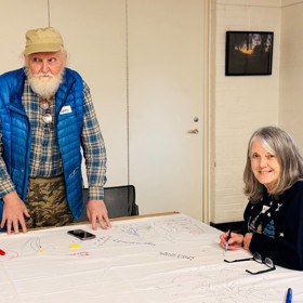 An older man and woman in colourful clothes, smiling - he stands, she sits drawing at a table.