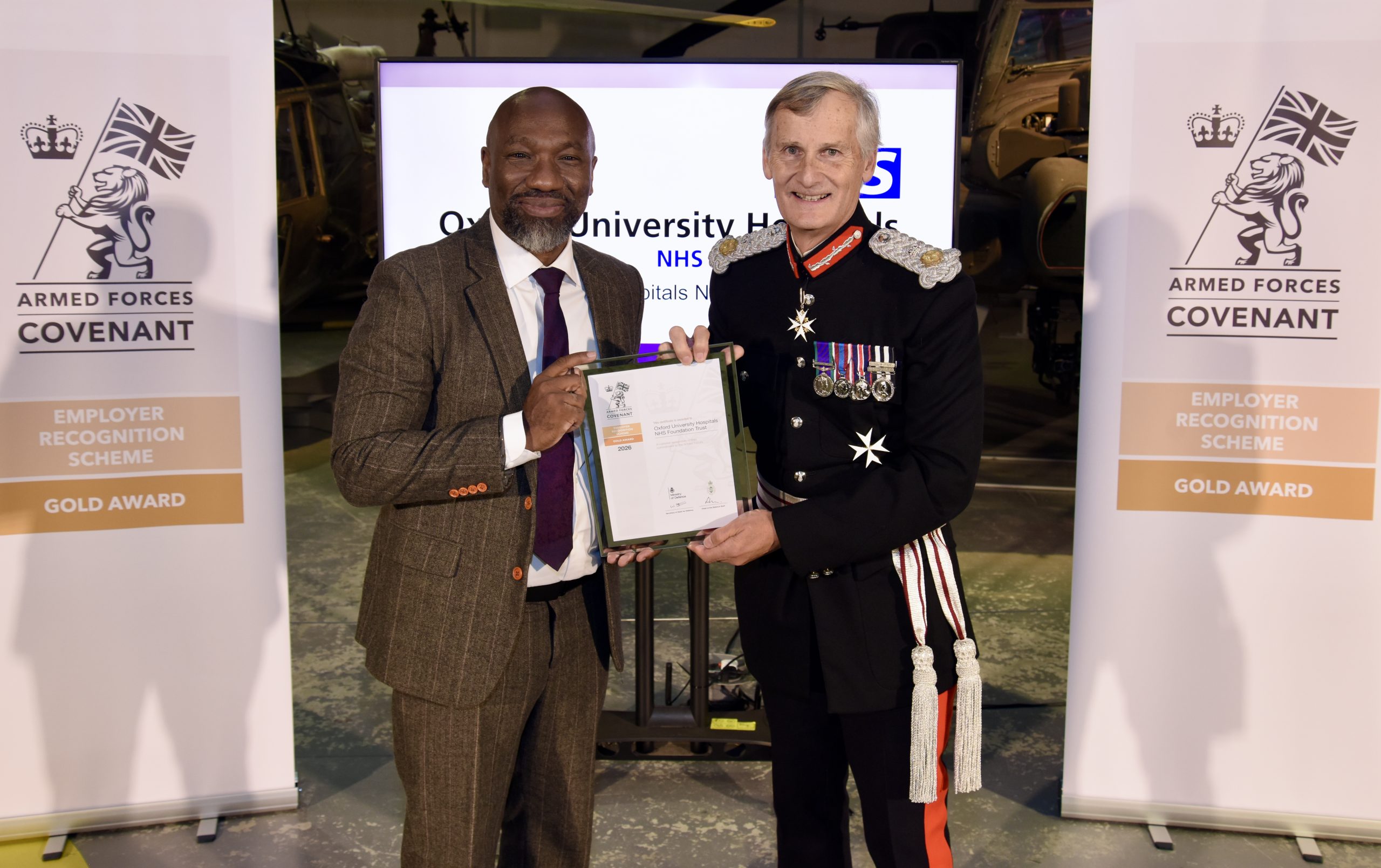 Two people holding OUH’s Gold Award certificate between Armed Forces Covenant banners. In the background in the OUH logo