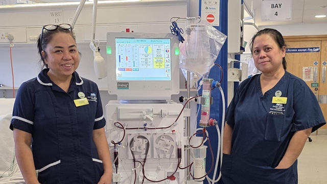 Two healthcare staff in clinical uniforms standing beside a dialysis machine in a hospital ward area.