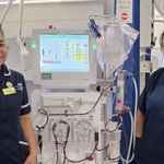 Two healthcare staff in clinical uniforms standing beside a dialysis machine in a hospital ward area.