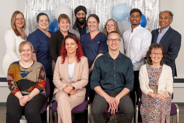 12 smiling people, some in healthcare uniform, arranged in front of a festive curtain and balloons