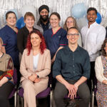 12 smiling people, some in healthcare uniform, arranged in front of a festive curtain and balloons