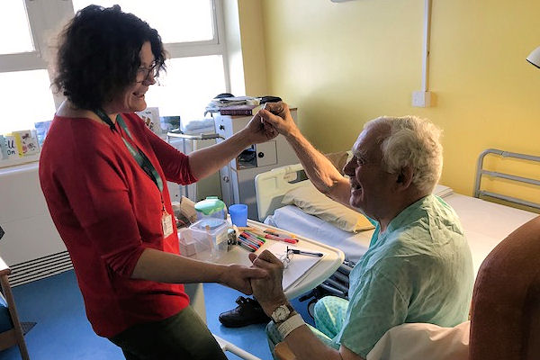 Woman in bright T-shirt and lanyard smiles and takes the hands of elderly seated man in hospital room.