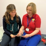 Two people sat down, smiling. A virtual reality headset is next to them. Laura, right, is wearing the red Play team t-shirt and is speaking with a teenage patient