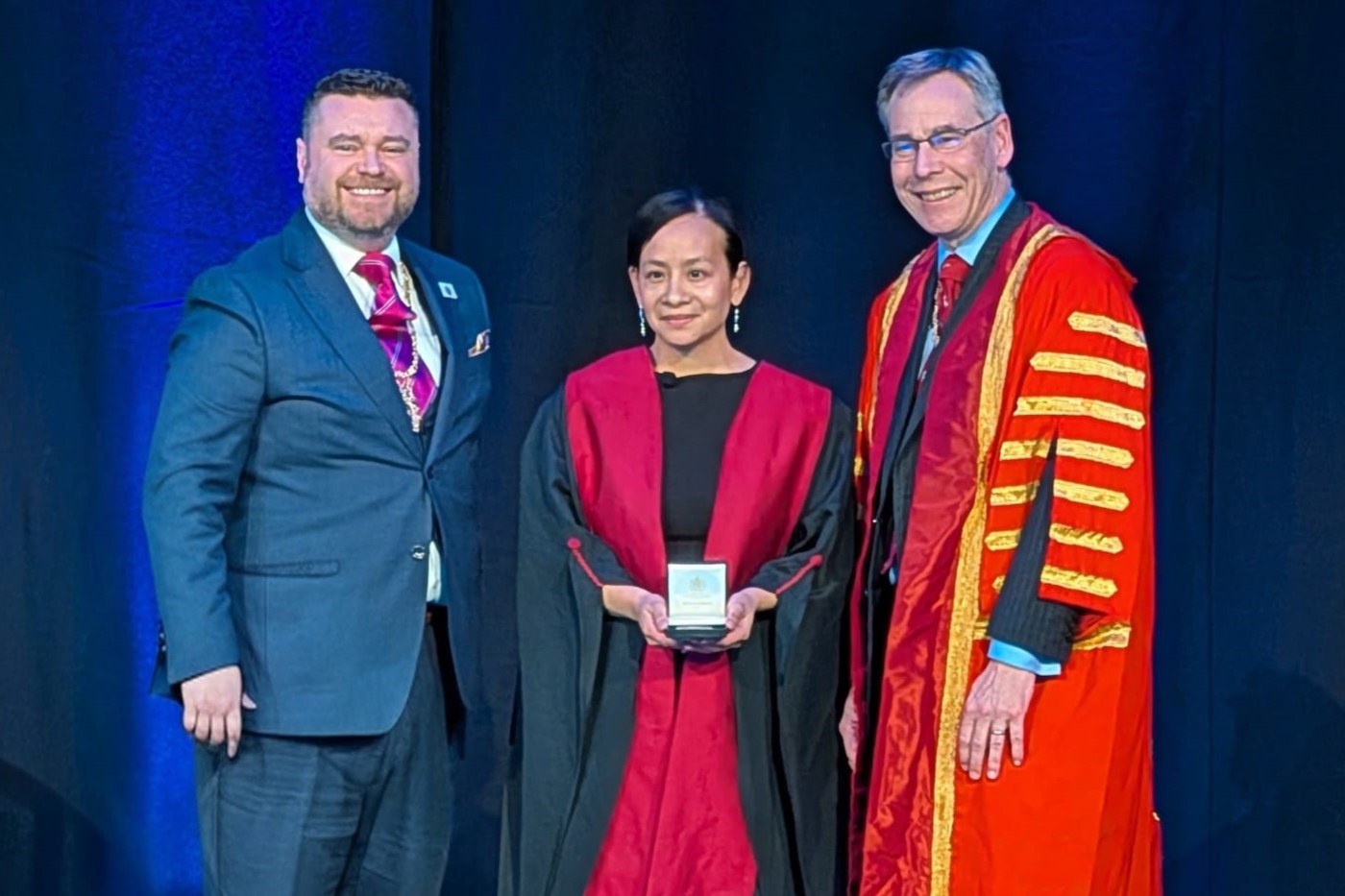 Three people pose on a stage; the centre person in a red academic gown holds the award, flanked by two others in formal and ceremonial attire.