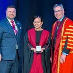 Three people pose on a stage; the centre person in a red academic gown holds the award, flanked by two others in formal and ceremonial attire.