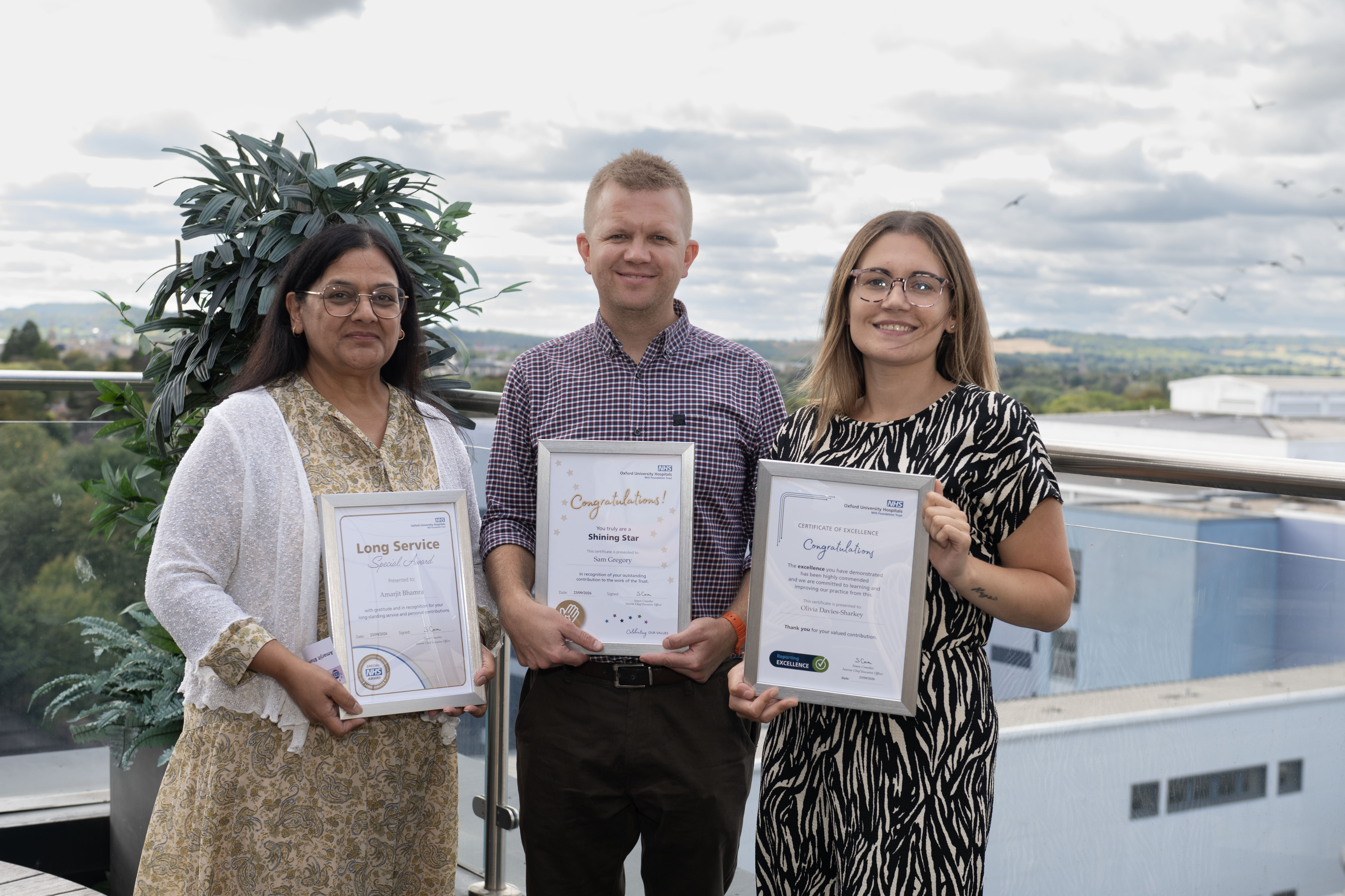 Three people standing outdoors, each holding a framed staff recognition certificate