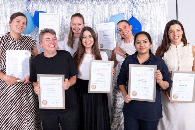 Seven smiling women, in summer clothes, holding framed certificates, in front of a sparkling backdrop.