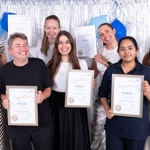 Seven smiling women, in summer clothes, holding framed certificates, in front of a sparkling backdrop.