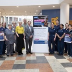 A group of healthcare staff standing in a hospital atrium beside a large “Martha’s Rule” information banner, with some team members holding promotional posters.