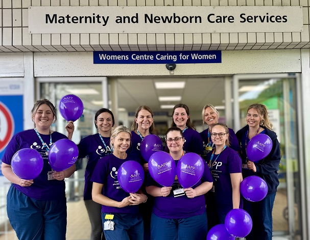 Nine healthcare workers in purple shirts hold matching balloons outside a “Maternity and Newborn Care Services” entrance.