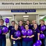Nine healthcare workers in purple shirts hold matching balloons outside a “Maternity and Newborn Care Services” entrance.