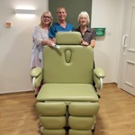 Three people behind a specialist green medical chair in a room with wooden flooring.