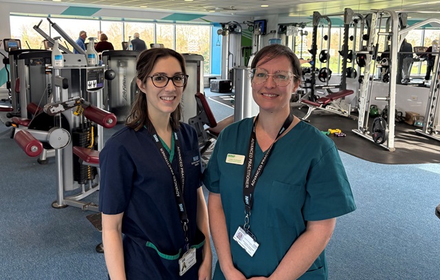 Two healthcare staff in clinical uniforms standing inside a gym, with exercise machines and weights visible in the background.
