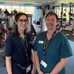 Two healthcare staff in clinical uniforms standing inside a gym, with exercise machines and weights visible in the background.