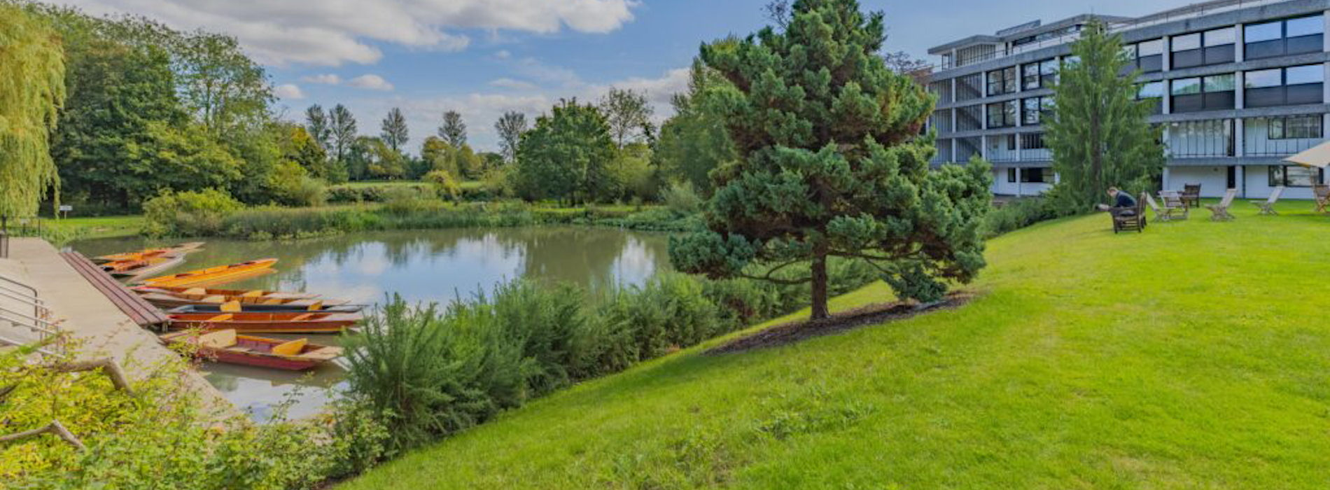 Wolfson College garden with lake, punts, green slope, tree and modern building in background; blue sky with white clouds.