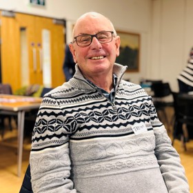 A seated smiling older man with the name sticker 'Trevor' on his patterned jumper.