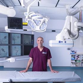 Man in burgundy T-shirt stands in clean hi-tech clinical setting with large machines around him, resting his hands on a scanner bed in front of him.