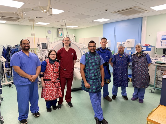 A group of seven clinical staff wearing scrubs and aprons standing together in a CT intervention suite, with medical equipment and monitors visible around them