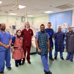 A group of seven clinical staff wearing scrubs and aprons standing together in a CT intervention suite, with medical equipment and monitors visible around them