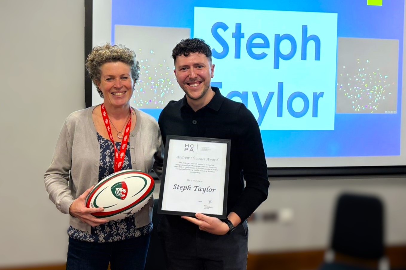 Two people stand in front of a presentation screen announcing “Steph Taylor” as the winner, with one holding a rugby ball and the other holding an award certificate.