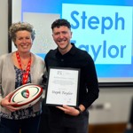 Two people stand in front of a presentation screen announcing “Steph Taylor” as the winner, with one holding a rugby ball and the other holding an award certificate.