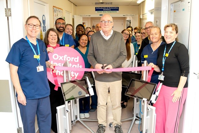 Group of nearly 20 people, some wearing clinical uniform. A patient in the middle prepares to cut a pink ribbon. There is a banner that reads "Oxford Hospitals Charity"