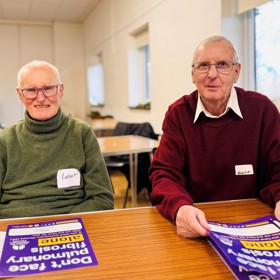 Two older men with name stickers on their jumpers, seated at a long table, holding posters that read 'Don't face pulmonary fibrosis alone'.