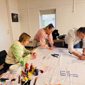 A man and two women in colourful shirts write on a table covered with a large sheet of paper - pens, paints and stickers in foreground.