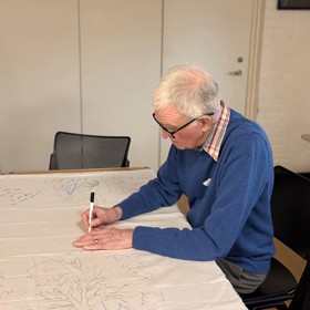 An older man in a jumper carefully draws lungs in felt tip on the corner of a large sheet on a table.