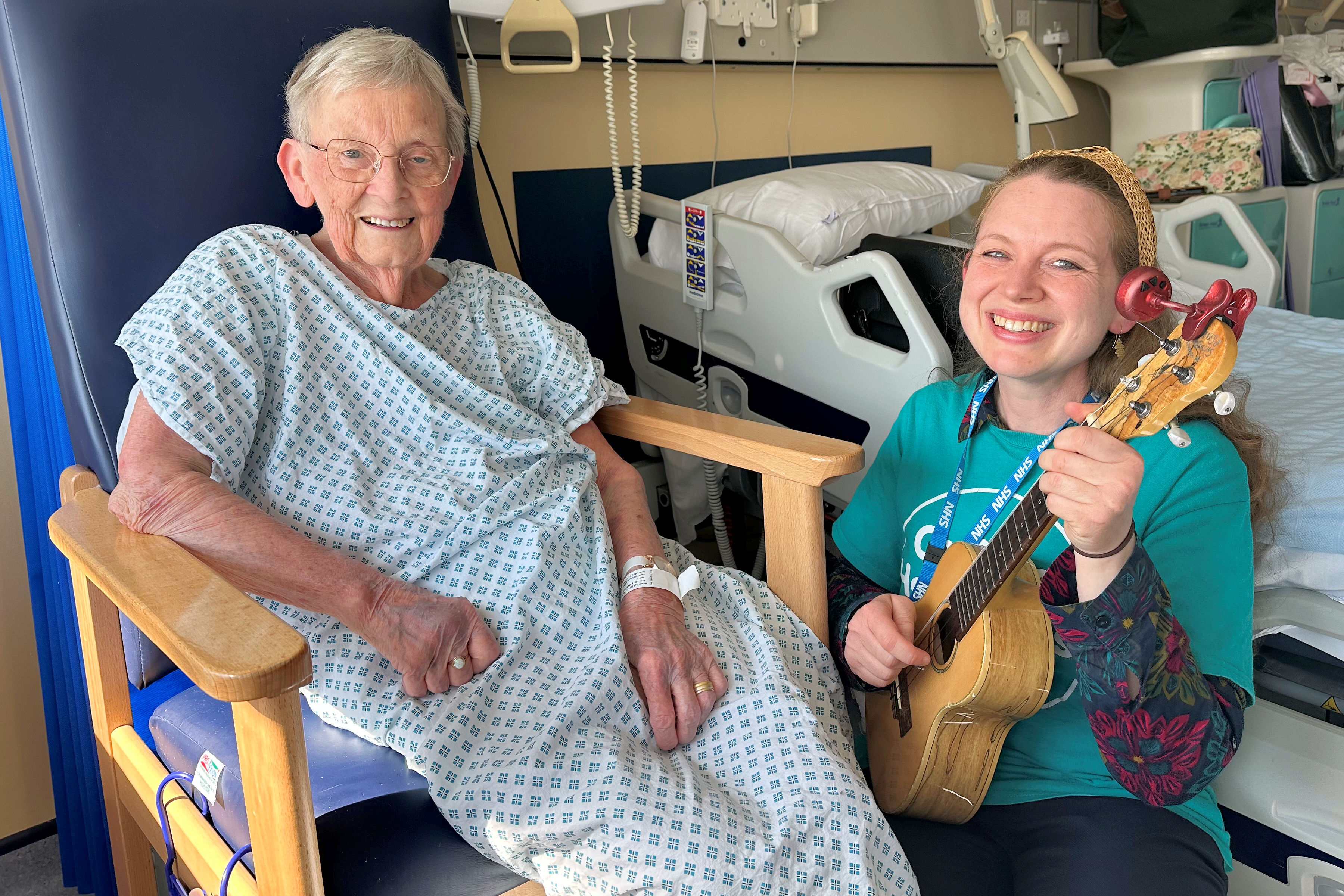 Smiling young woman with small guitar with seated elderly female patient in hospital room.