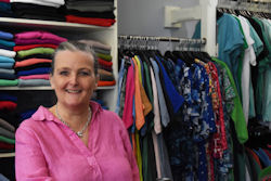 Smiling woman in bright shirt with shelves of colourful folded clothing and racks of dresses behnid her