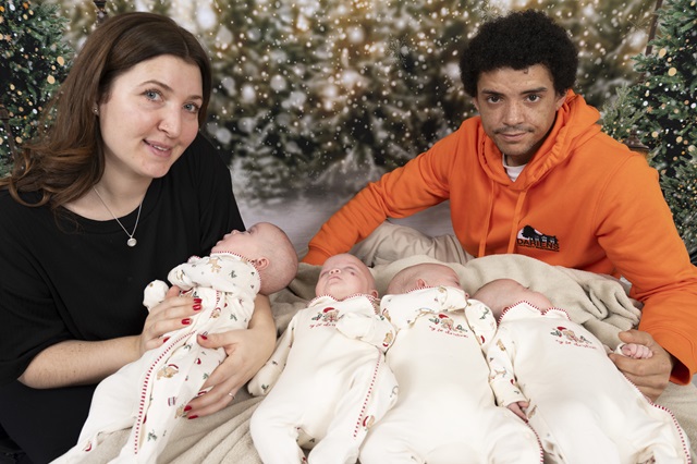 Mum and dad with their quadruplets, who are lying down and wearing matching Christmas outfits. There is a festive display in the background