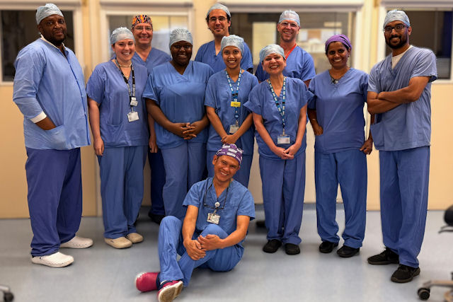 A group of 11 smiling people wearing theatre scrubs in a clinical area