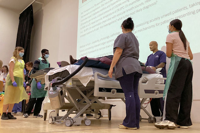 Five people in NHS uniform or colourful PPE stand on a stage around a volunteer in a hospital bed.