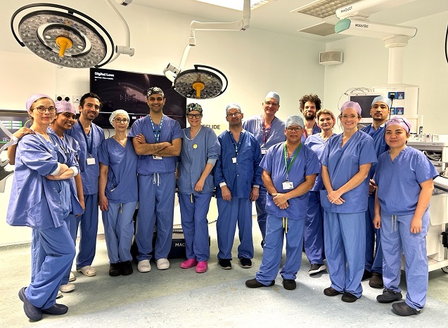 A group of medical professionals in blue surgical scrubs and caps stand together in a brightly lit operating room. Overhead surgical lights and medical equipment are visible.