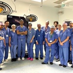 A group of medical professionals in blue surgical scrubs and caps stand together in a brightly lit operating room. Overhead surgical lights and medical equipment are visible.
