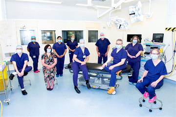 A group of 10 uniformed staff in surgical masks, seated apart in an operating theatre