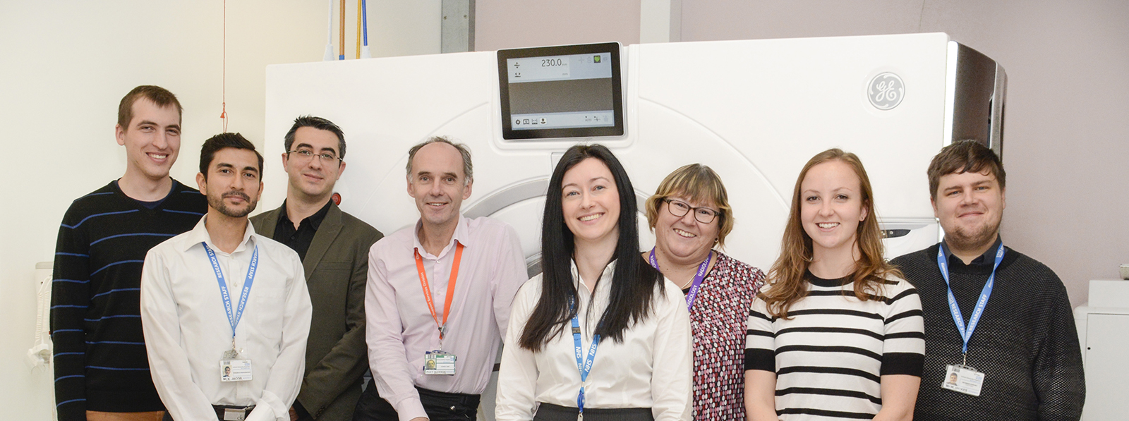Eight smiling people with lanyards in front of an MRI scanner