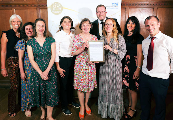 Nine smiling smartly dressed people display a certificate in a panelled room