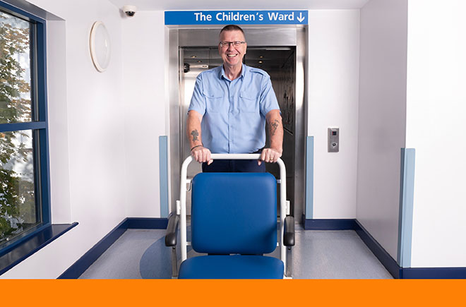 Smiling man in shirt sleeves pushes empty wheelchair out of lift door under sign saying 'The Children's Ward'